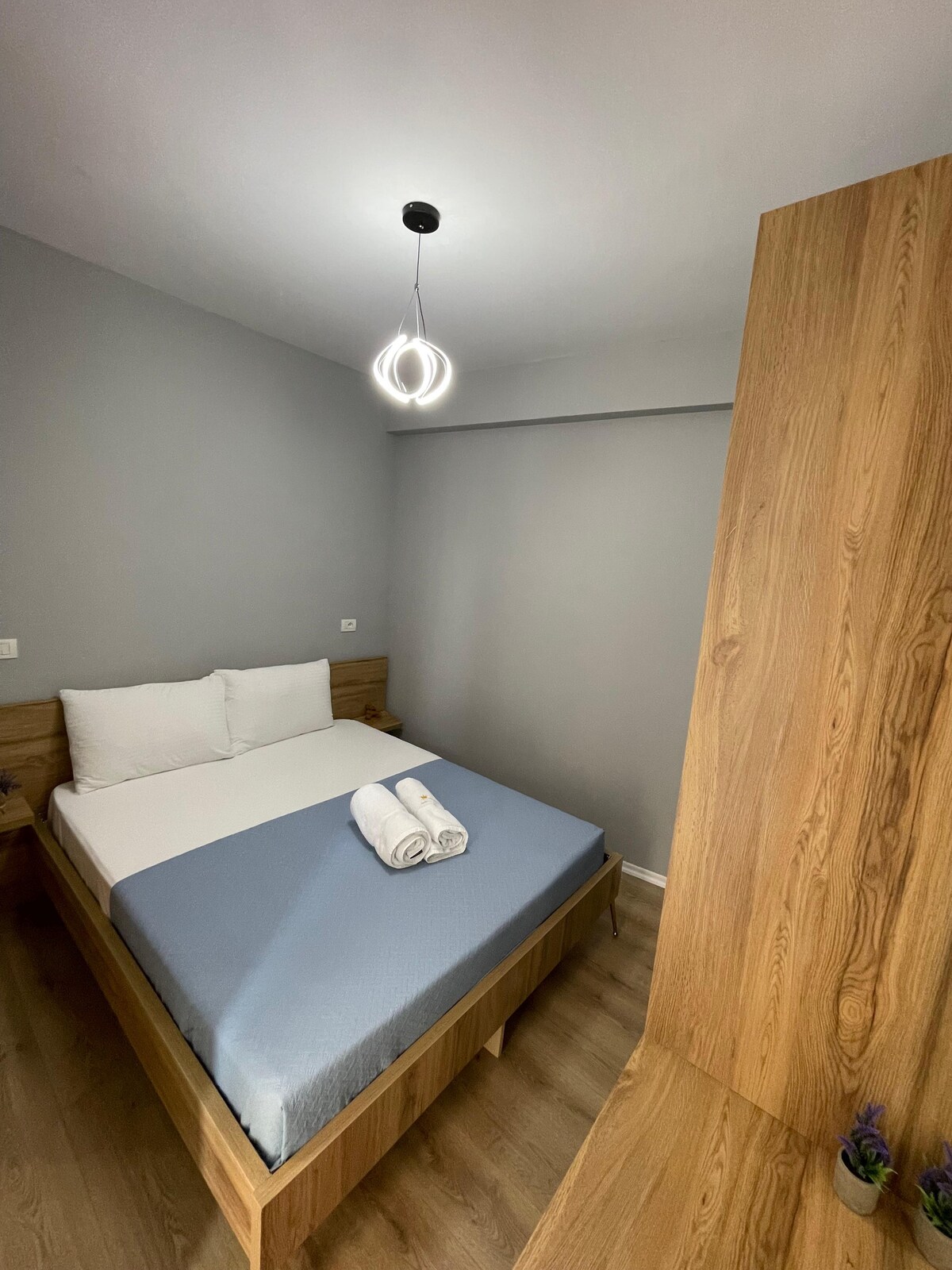 A cozy sleeping space is presented with a light-colored wooden bed featuring a soft blue bedspread. Fluffy white towels are neatly arranged on the bed. A modern light fixture hangs from the ceiling, and neutral grey walls contribute to a serene ambiance.