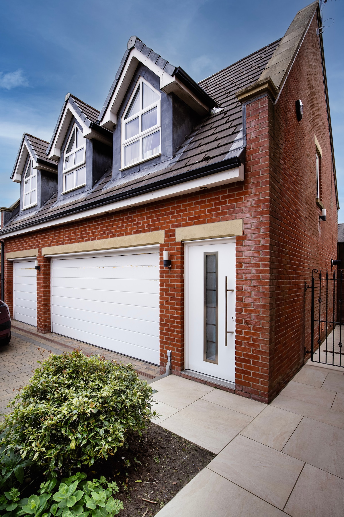 The exterior showcases a modern brick facade with three prominent gable windows. A sleek white front door stands at the entrance, complemented by a porch area and neatly arranged pavers. Lush greenery is present in the foreground, enhancing the inviting appearance of this coastal escape.