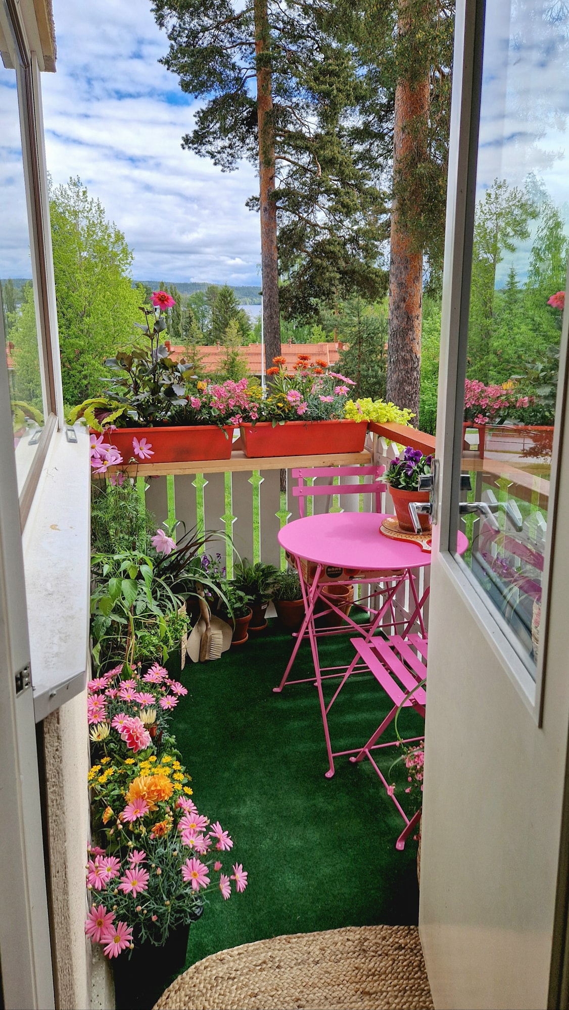 A small balcony is visually accessible through an open door, featuring a pink table and chairs surrounded by a variety of flowers. Lush green plants are arranged along the railing, creating a vibrant, inviting space. A bright area rug complements the greenery.