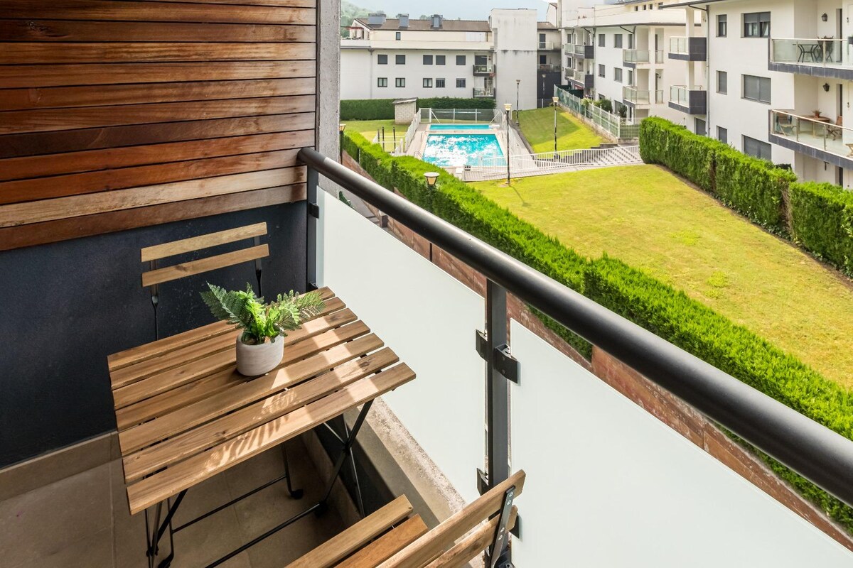 A balcony area features a wooden table and folding chairs, accompanied by a small potted plant. The view includes a well-maintained green lawn and a community swimming pool, framed by modern apartment buildings in the background.