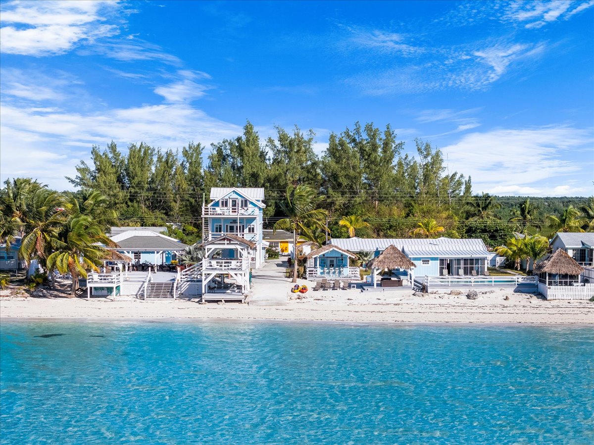 A beachfront property is seen from above, featuring multiple structures nestled among palm trees. Sandy paths lead from the beach to wooden decks, while the crystal-clear turquoise water gently laps at the shore. The vibrant scenery reflects a tranquil island atmosphere.