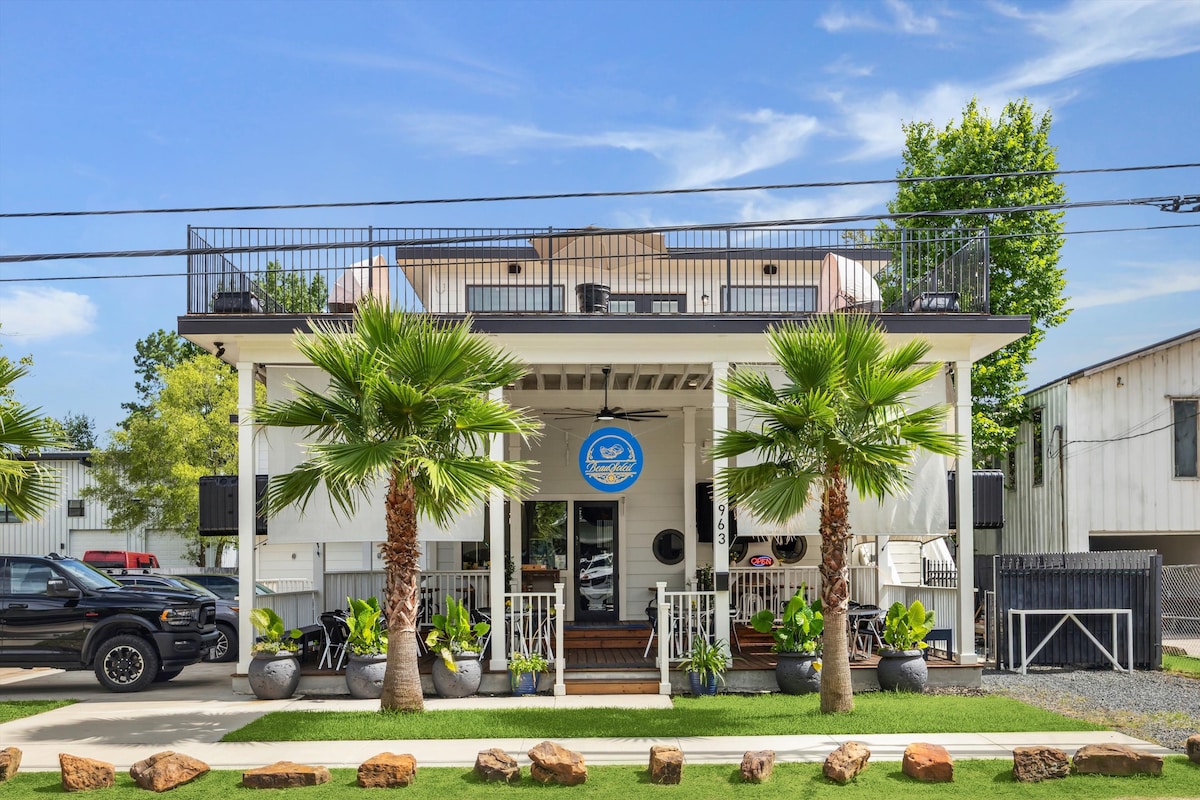 A two-story building features a welcoming entrance, surrounded by lush greenery and palm trees. A spacious rooftop terrace is seen above, offering an open space adorned with railings. The inviting façade is complemented by large windows and a bright blue sign, signifying the property's name.