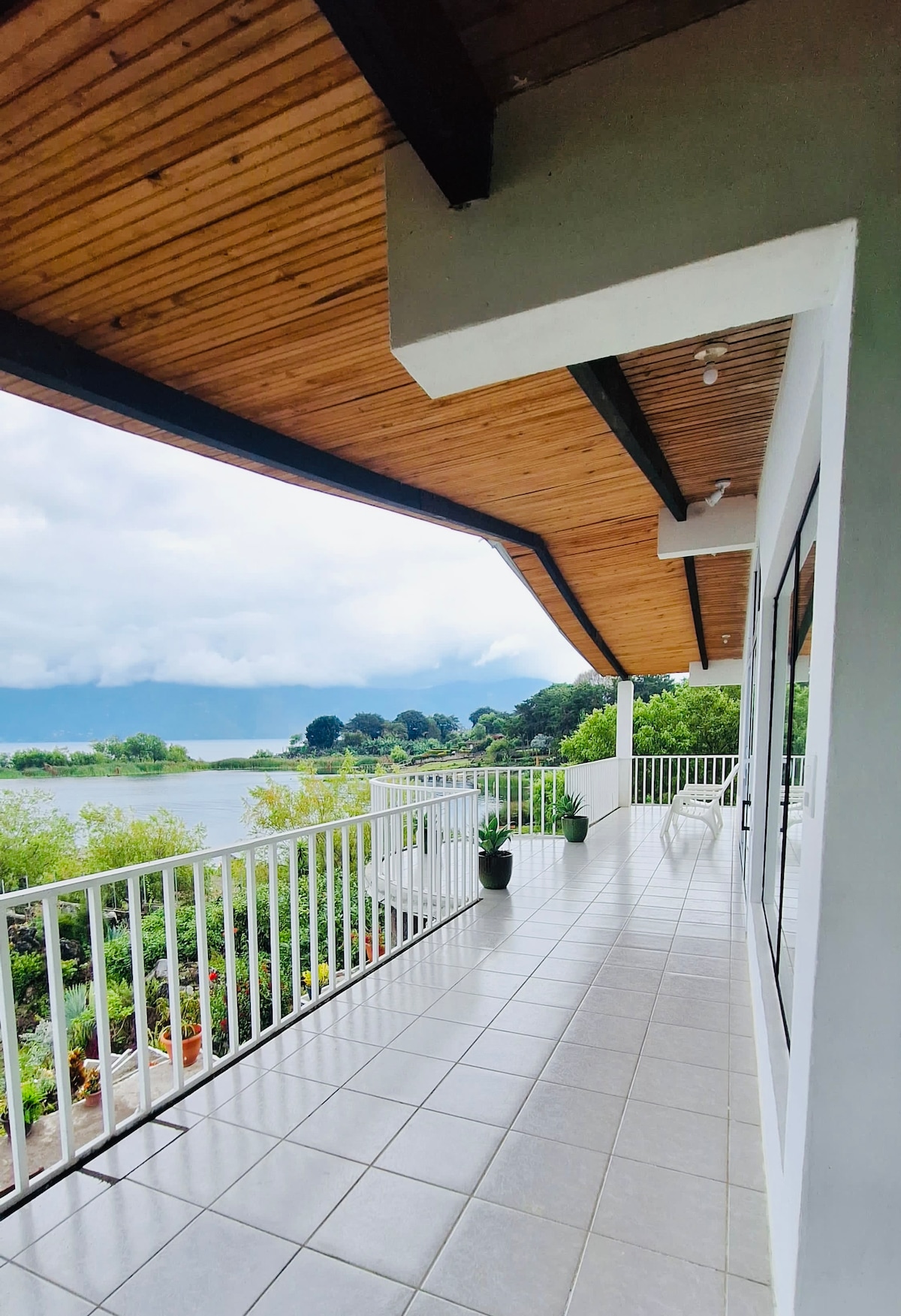 A spacious balcony extends along the edge of the villa, featuring a white railing and tiled flooring. Potted plants add greenery to the space, offering views of Lake Atitlan and the lush surroundings under a partially cloudy sky.