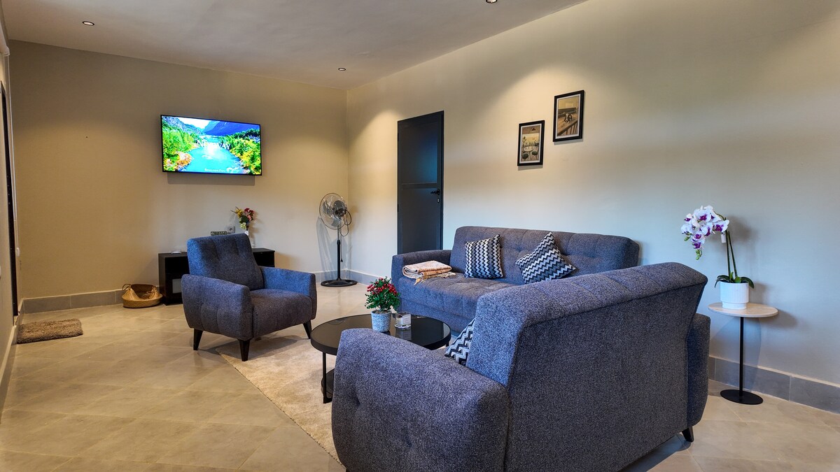 A cozy living area is highlighted by a comfortable gray sofa and two matching armchairs arranged around a dark circular coffee table. A flat-screen television is mounted on the wall, while a small fan stands beside a potted orchid, adding a touch of greenery.
