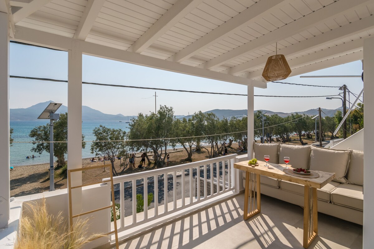 A balcony is presented with a modern seating area, featuring beige cushions and a low table set with refreshments. The panoramic view of the beach and sea is clearly visible, with soft waves lapping at the shore under a bright sky.