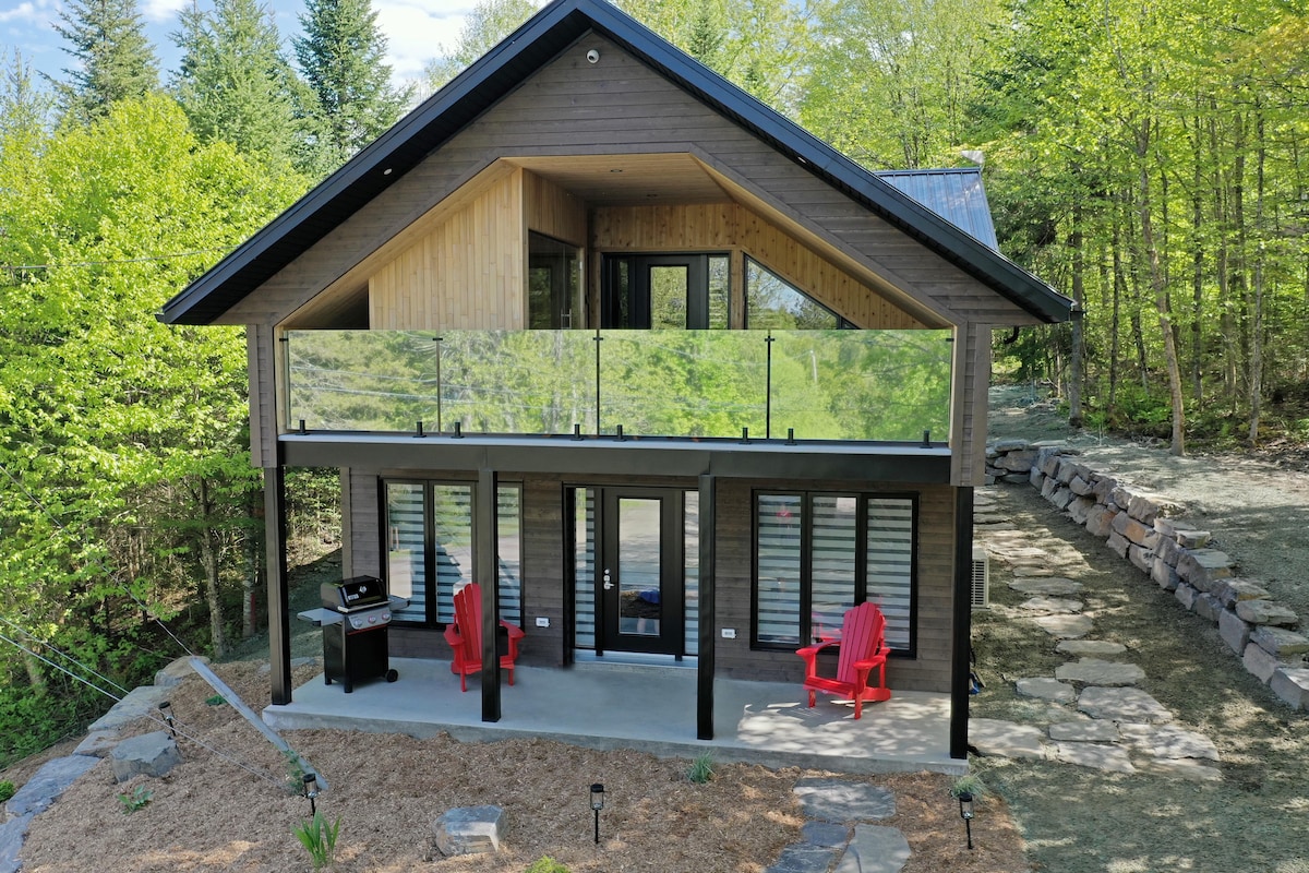 A contemporary cabin is surrounded by lush greenery, featuring a spacious deck with glass railings. Two red Adirondack chairs are positioned on a gravel patio, with a grill nearby. Large windows allow for ample natural light, creating a welcoming outdoor space.