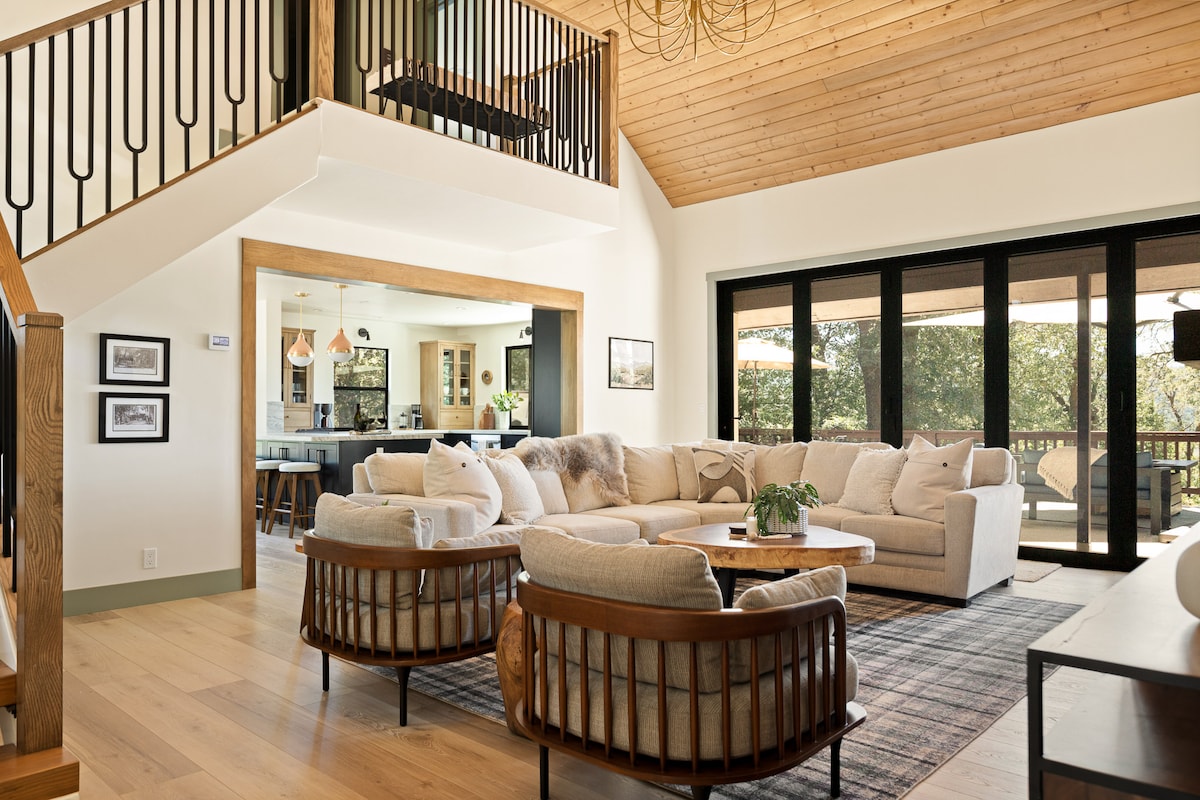 A spacious living area features an inviting sectional sofa accompanied by two uniquely designed armchairs. Natural light streams through large doors that connect to the outdoor deck, while warm wooden accents and a ceiling illuminate the modern mountain design.