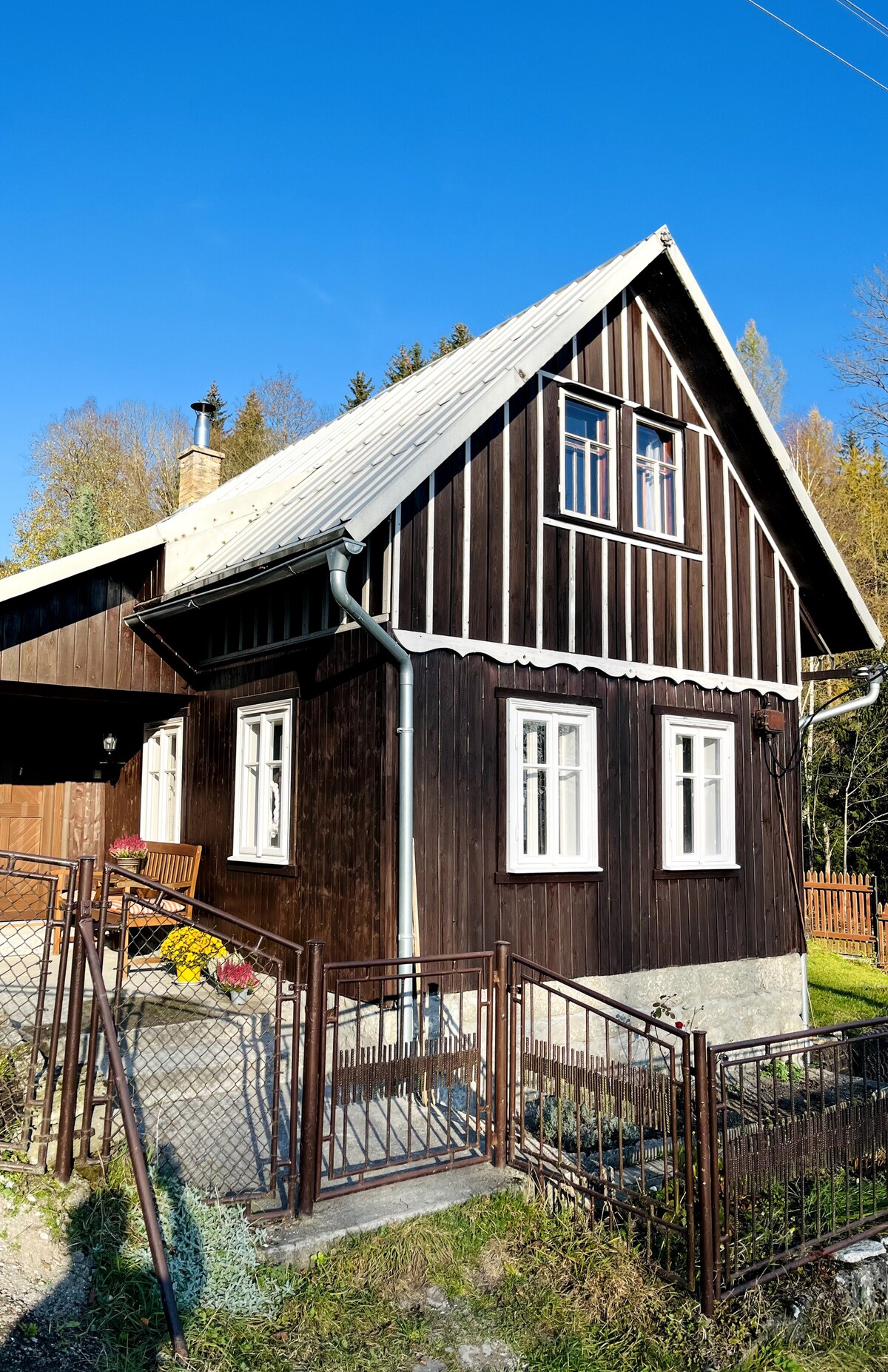 A charming wooden cottage features a sloped roof and a dark exterior, complemented by light-colored window frames. The front yard is enclosed with a metal fence. Seasonal flowers are placed near the entrance, surrounded by a peaceful natural setting.