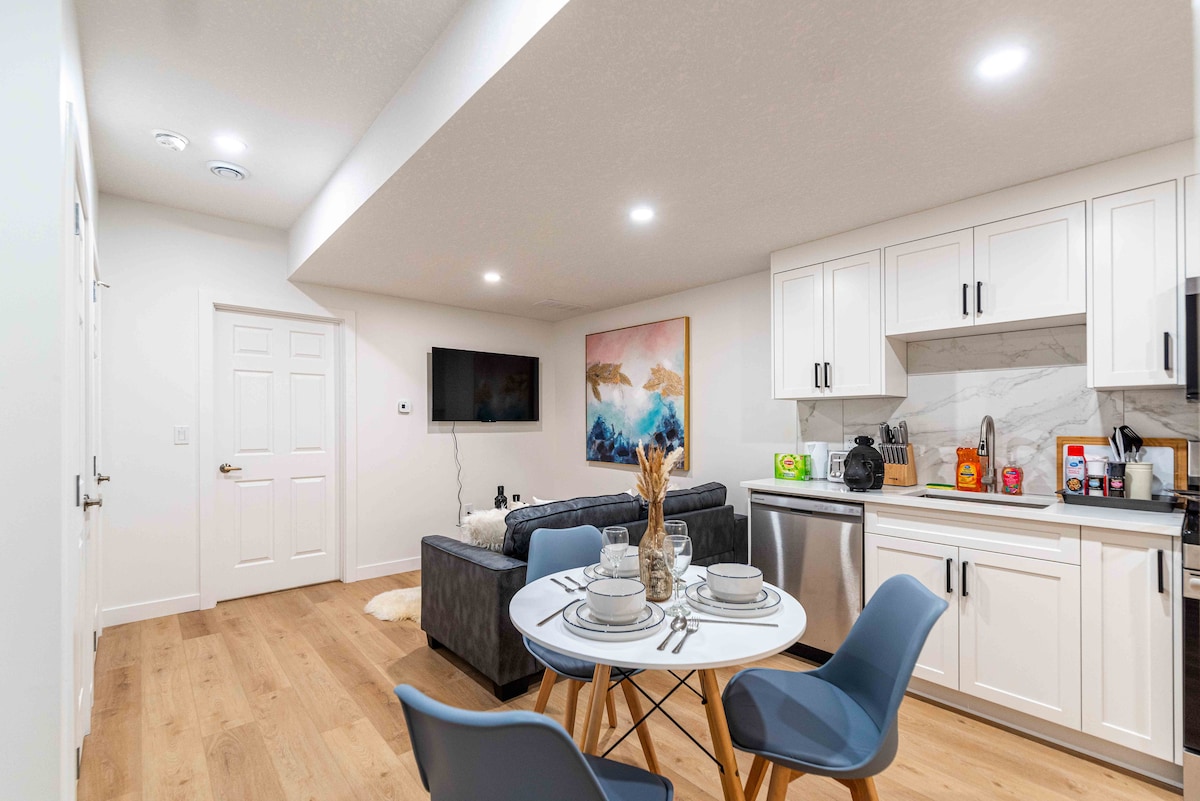 An open living space is featured, highlighting a modern kitchen with white cabinetry and stainless steel appliances. A dining table is set for four, accompanied by a comfortable seating area with a dark sofa. A wall-mounted TV and a decorative wall art piece enhance the room.