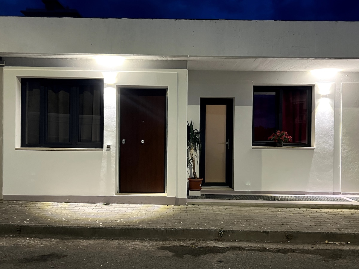 The exterior of the recently renovated property is shown, featuring two separate entrances. Soft lighting illuminates the clean white walls, enhancing the inviting appearance. A potted plant is positioned near the entrance to the right, and large windows are visible, suggesting a comfortable atmosphere.