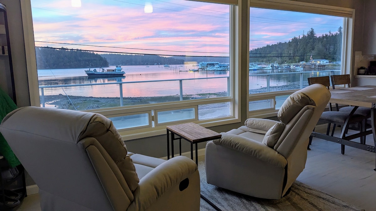 Two recliner chairs are positioned by large windows, offering a view of the serene waterfront at sunset. The sky is painted in soft hues of pink and purple, reflecting on the calm water. A small wooden table rests between the chairs.