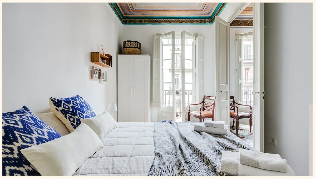 A spacious bedroom features a light color scheme with two large beds adorned with patterned blue and white bedding. Natural light floods the room through tall windows, which open to a balcony. Two elegant armchairs and a wardrobe are also visible.