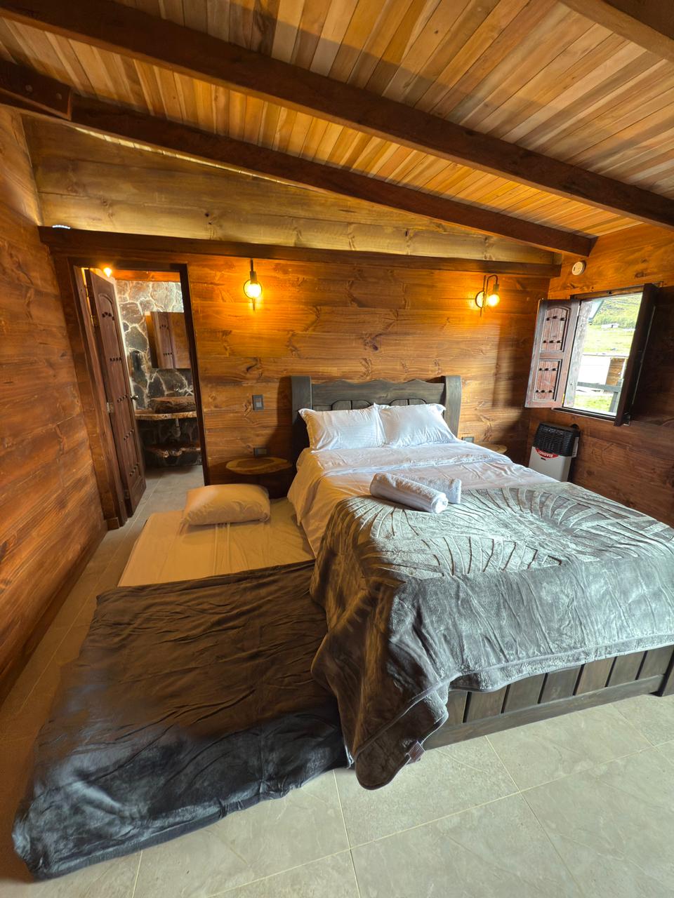 A rustic cabin room features a spacious bed covered with a soft gray blanket. Warm wooden walls are complemented by subtle lighting fixtures. A doorway leads to a private bathroom area, while natural light enters through the adjacent window, enhancing the tranquil atmosphere.
