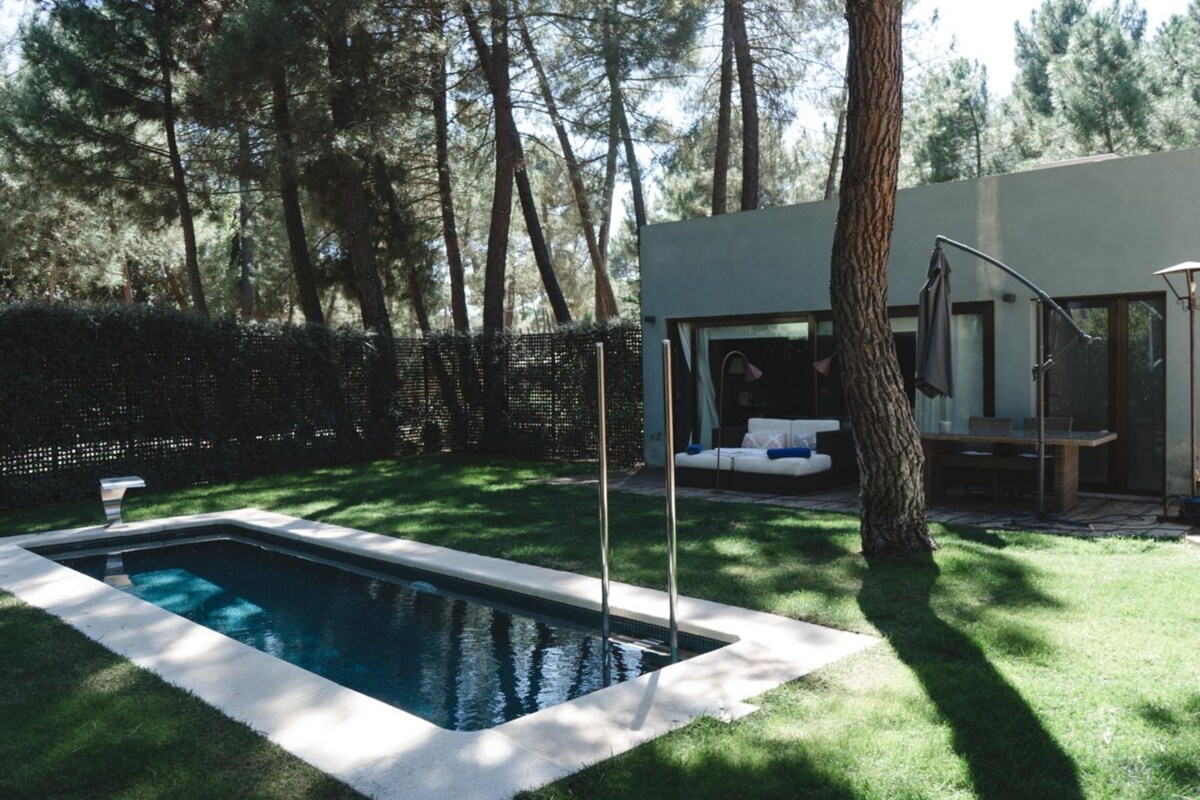 A private salt-water pool is set amidst lush green grass, surrounded by tall trees. The pool area features a shaded lounge with comfortable seating and an outdoor dining table, creating an inviting space for relaxation.