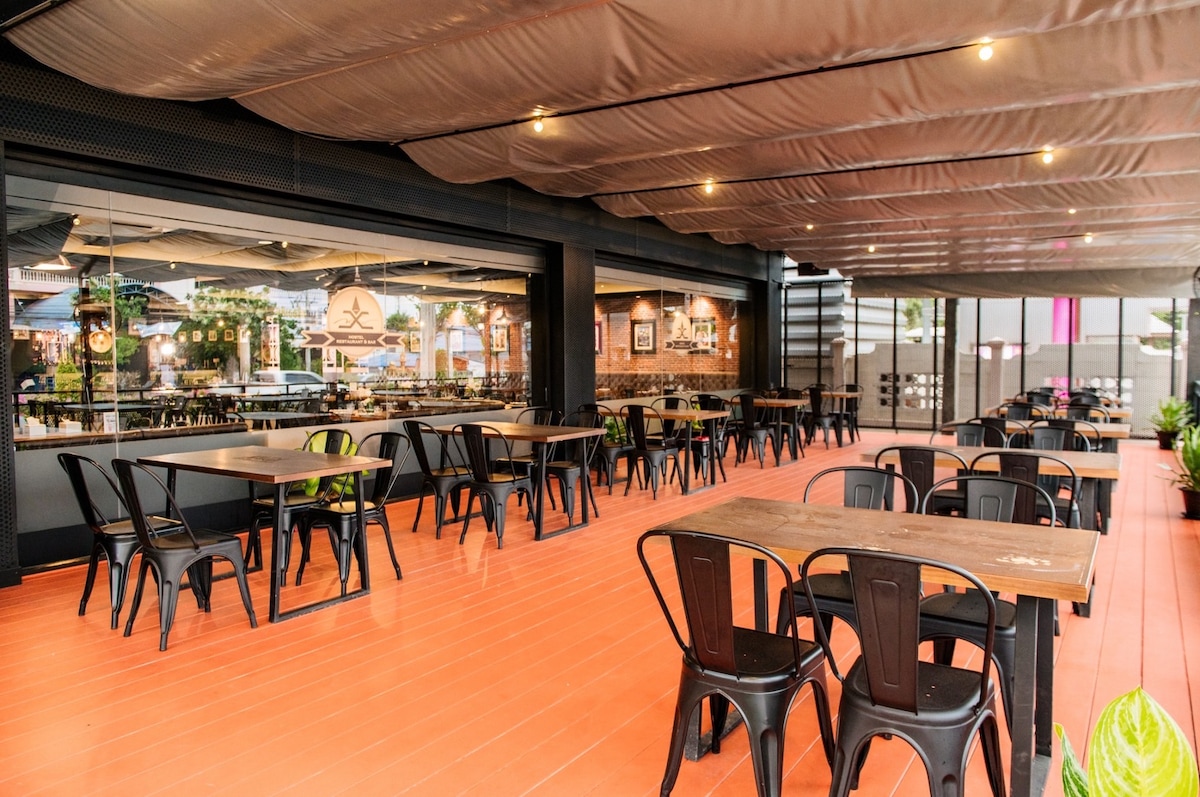 An open dining area features multiple wooden tables surrounded by black metal chairs. Large glass windows provide a view of the outdoors, while overhead lighting and a shaded ceiling create a comfortable environment for guests.