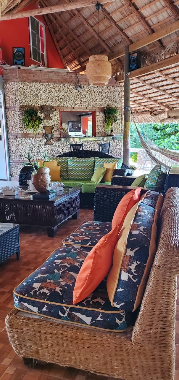 An open, shaded living area features woven furniture with colorful cushions. A dark wooden coffee table is centered among the seating, while vibrant greenery is visible in the background. Natural materials, such as thatched roofing and stone accents, contribute to a warm environment.