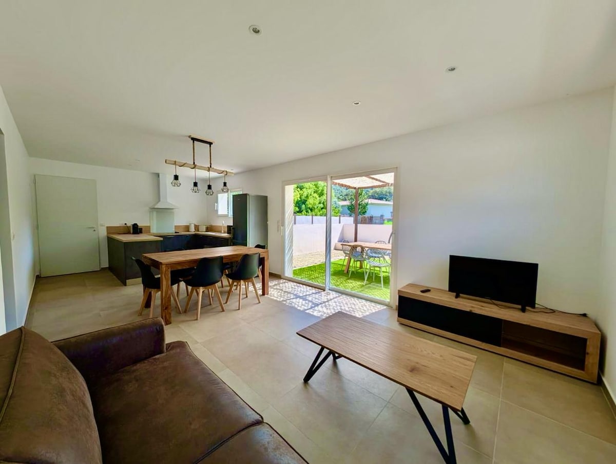 A modern living space is depicted, featuring an open layout with a comfortable seating area that includes a sofa. A dining table with chairs is visible, along with a kitchenette. Large sliding doors provide access to an outdoor area, enhancing natural light in the space.
