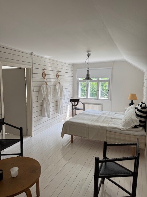 A light-filled bedroom features a spacious bed adorned with simple linens, complemented by two hanging robes on the wall. A window offers views of the greenery outside, while two black chairs and a round table provide seating options in the room.
