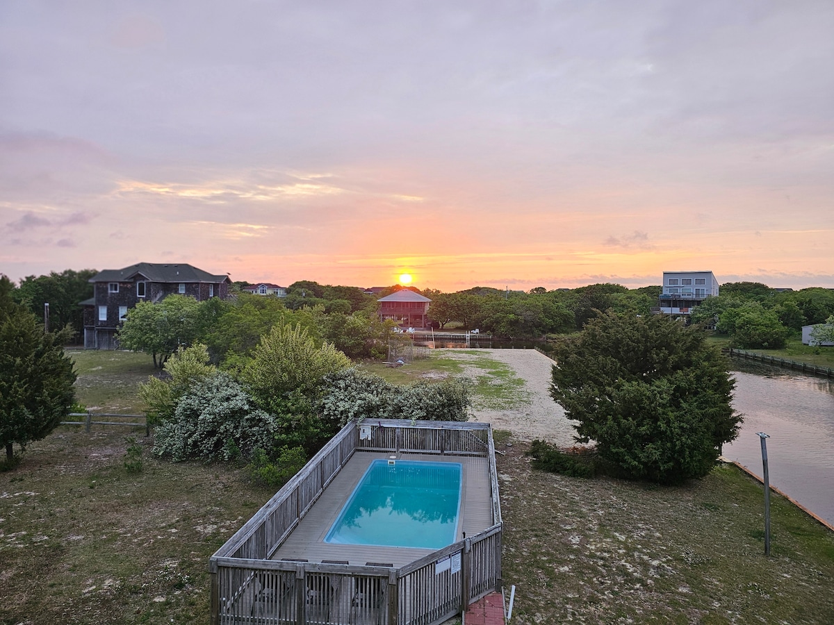 A private pool is situated on a sunken deck, reflecting the warm hues of the sunset. Lush greenery surrounds the area, creating a serene backdrop. The horizon features a mix of trees and nearby homes, contributing to a tranquil atmosphere.