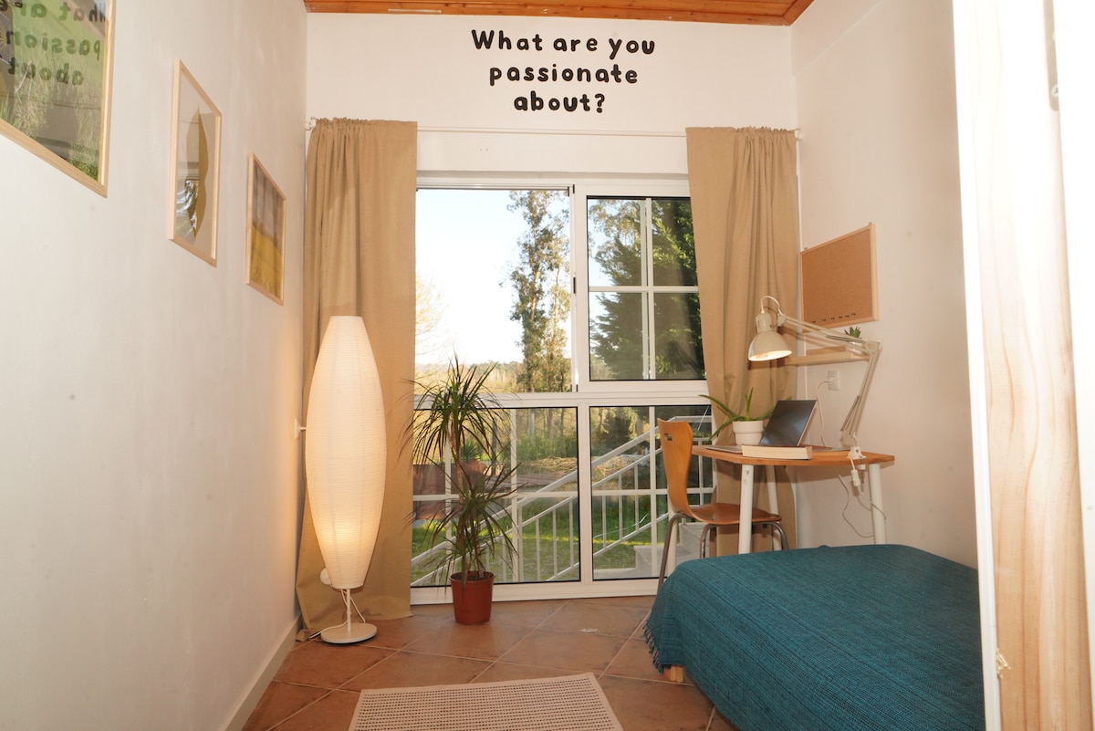 A cozy room features a spacious bed covered with a blue blanket. A desk with a lamp and a chair is positioned nearby, while a large window allows natural light in, revealing greenery outside. Inspirational text is displayed on the wall above.