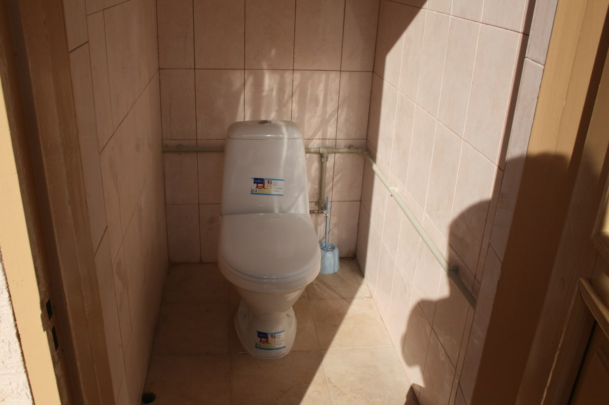A clean and simple restroom is depicted, featuring a modern toilet positioned against light tiles. The space is illuminated by natural light filtering through, highlighting the tiled flooring and plumbing fixtures along the wall.