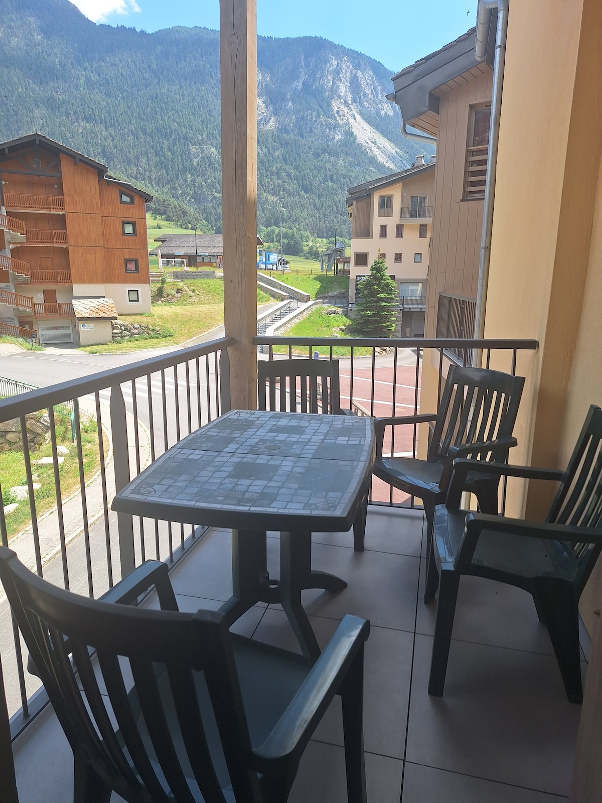 A balcony featuring a rectangular table with four black chairs is visible. The surrounding mountains create a scenic backdrop. The tile flooring and railing complement the outdoor space, providing a comfortable area for relaxation.