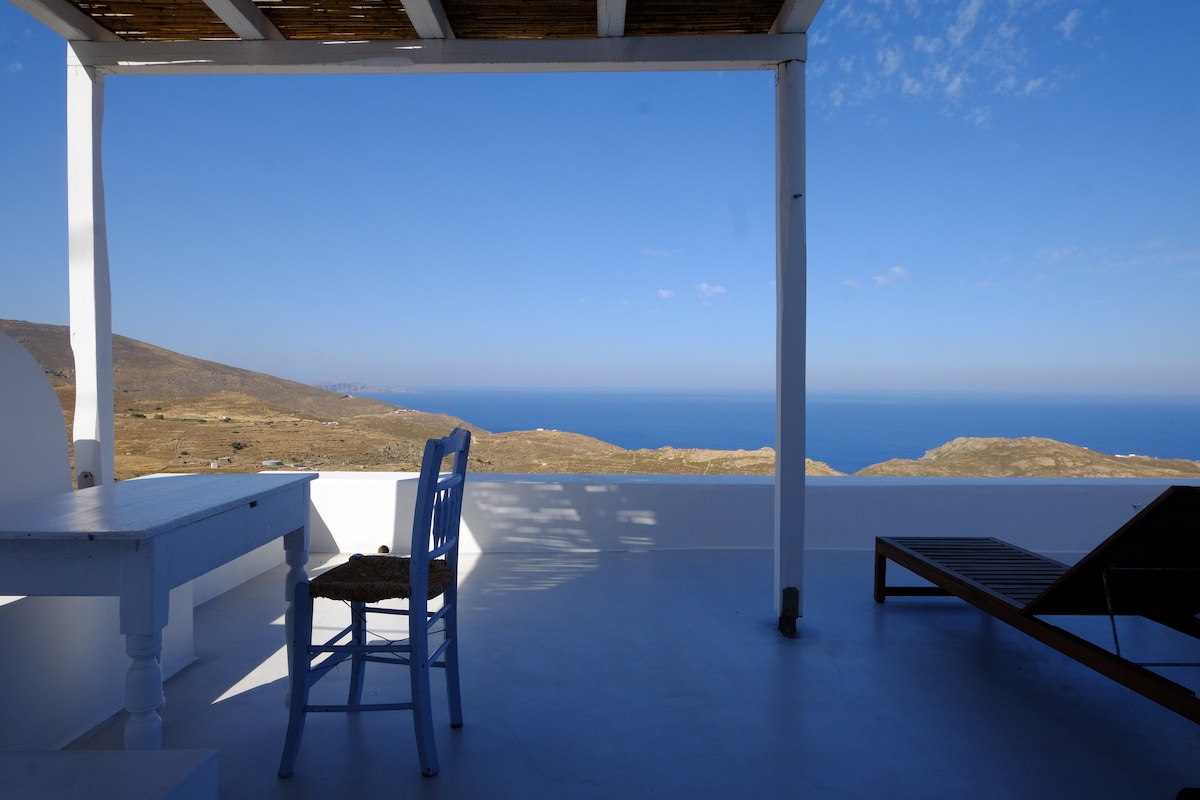 A spacious balcony features a simple white table and a traditional blue chair, complemented by a sun lounger. The panoramic view reveals the serene blue of the Aegean Sea and rugged landscapes, illuminated by a clear sky. The setting invites relaxation and appreciation of the scenery.