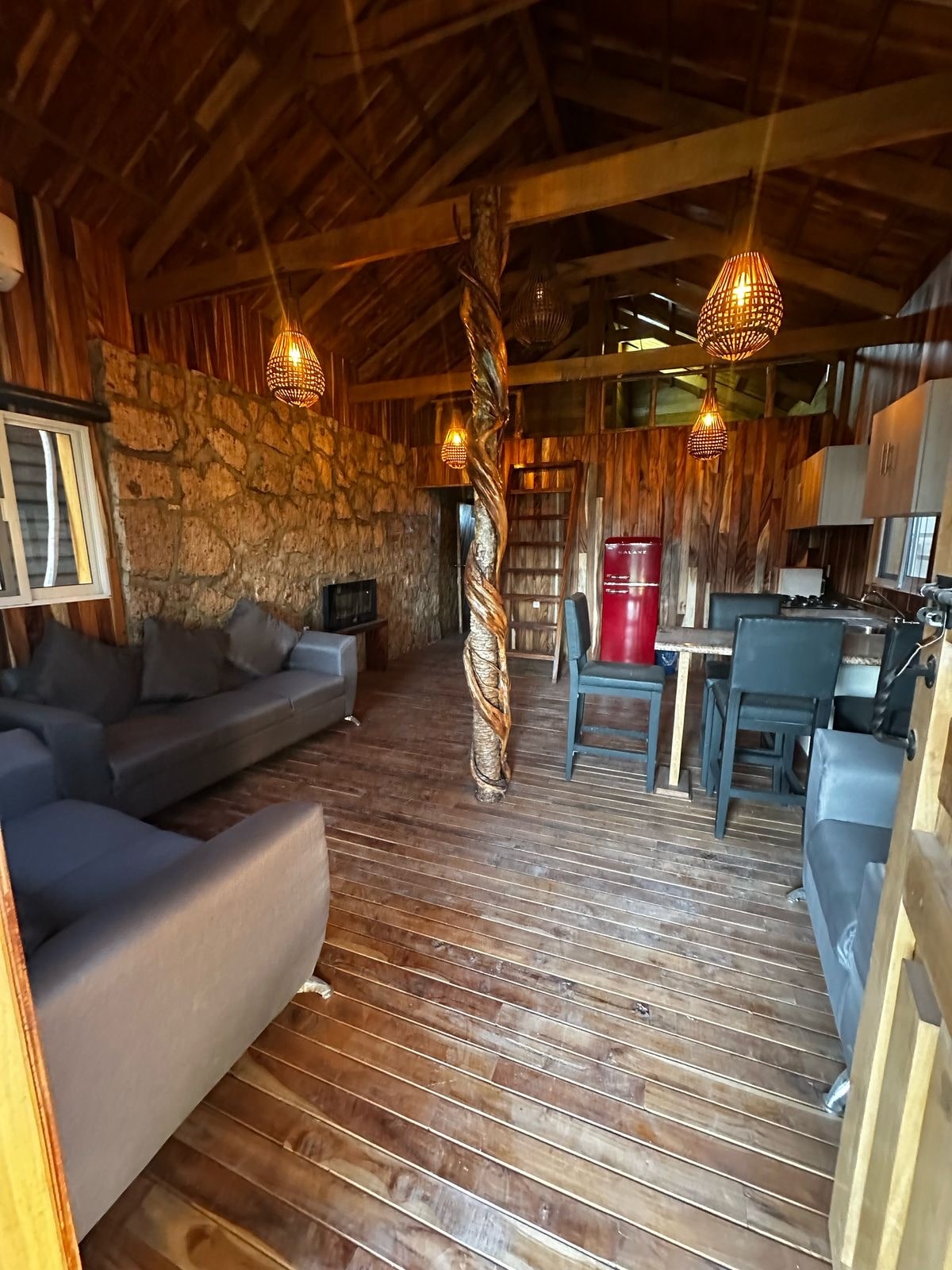 A cozy interior space features wooden walls and flooring, complemented by a stone accent wall. Soft seating is arranged around a central column. A dining table is positioned nearby, with light fixtures hanging above. A staircase leads to a loft area, adding height to the room.