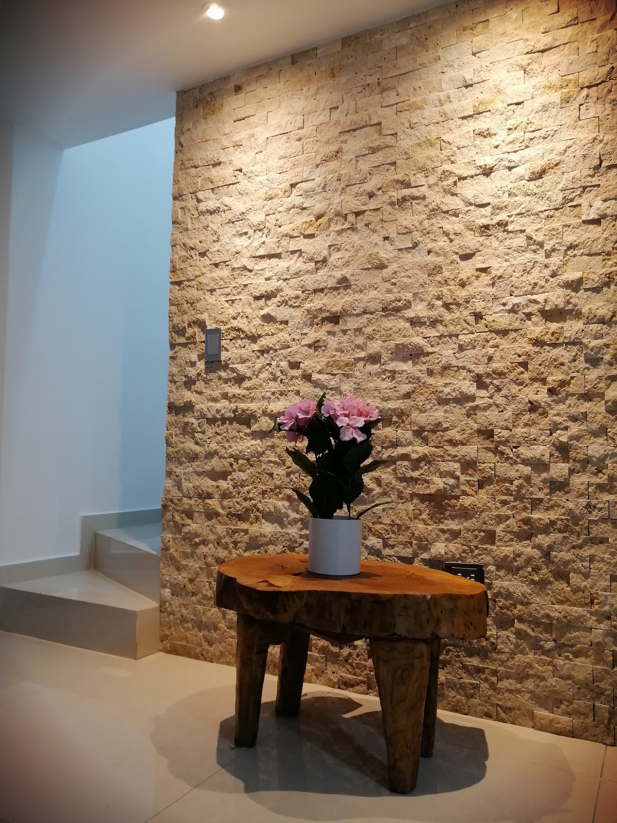 A warmly lit entryway features a textured stone wall, providing an inviting backdrop. A wooden table with a unique shape supports a potted arrangement of pink flowers. A staircase leading upward is visible in the background, enhancing the sense of space.