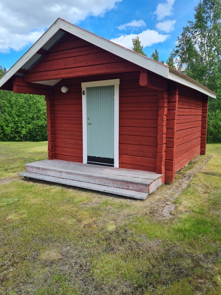 A red wooden cabin is situated on a grassy area, showcasing a simple design with a sloped roof. The front porch features unpainted wooden steps leading to a door with a light green panel. Surrounding greenery complements the rustic structure.