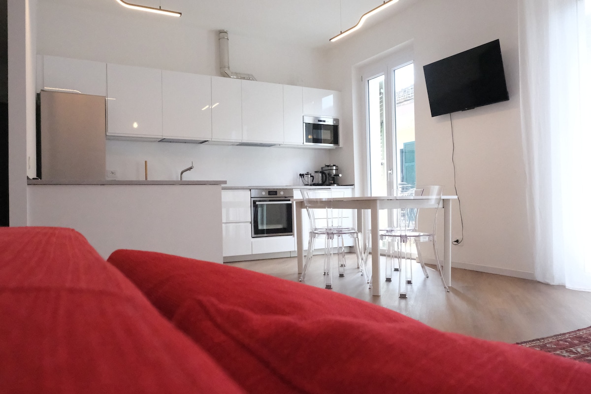 An open kitchen and dining area is visible, featuring sleek white cabinetry and modern appliances including an oven and microwave. A dining table with transparent chairs is positioned next to the kitchen. A red couch provides a contrasting element in the foreground, while a flat-screen TV is mounted on the wall.