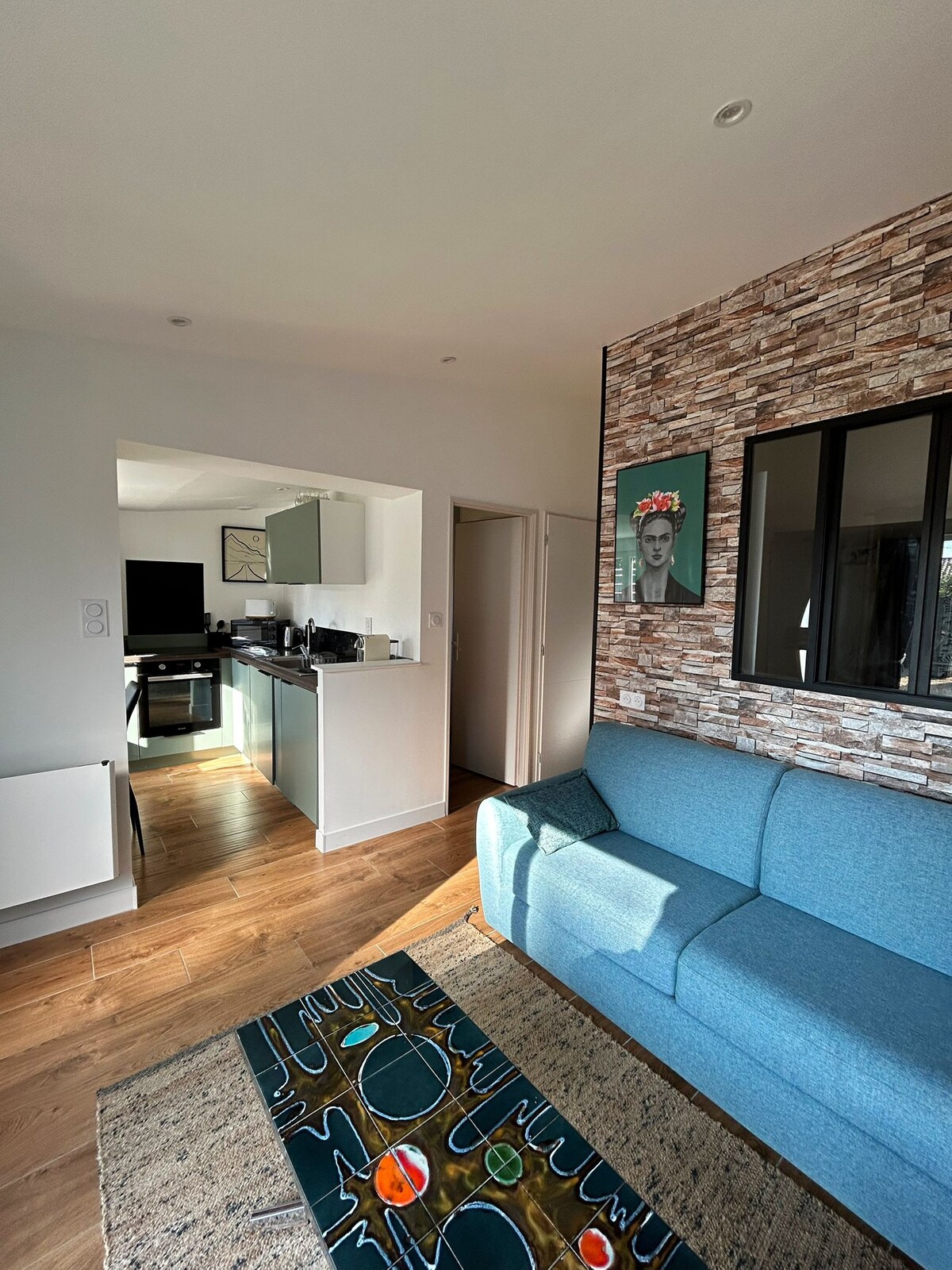 The living area features a blue sofa positioned next to a patterned area rug. A kitchenette is visible in the background, showcasing modern cabinetry and appliances. Natural light enters through a window, creating a bright and welcoming environment.