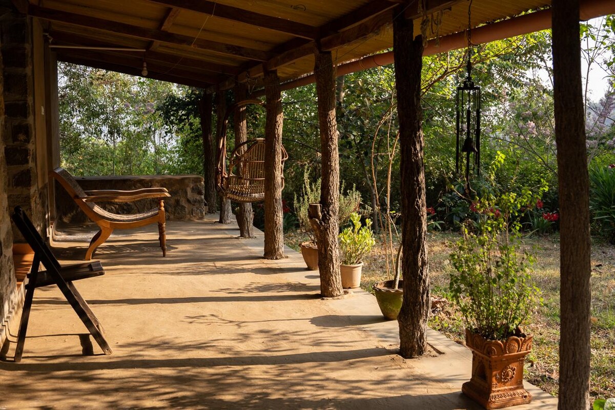 A covered porch is supported by wooden pillars, featuring a wooden swing and a couple of chairs. Potted plants and small shrubs add greenery to the space, with dappled sunlight creating patterns on the floor. The porch overlooks the surrounding natural landscape.
