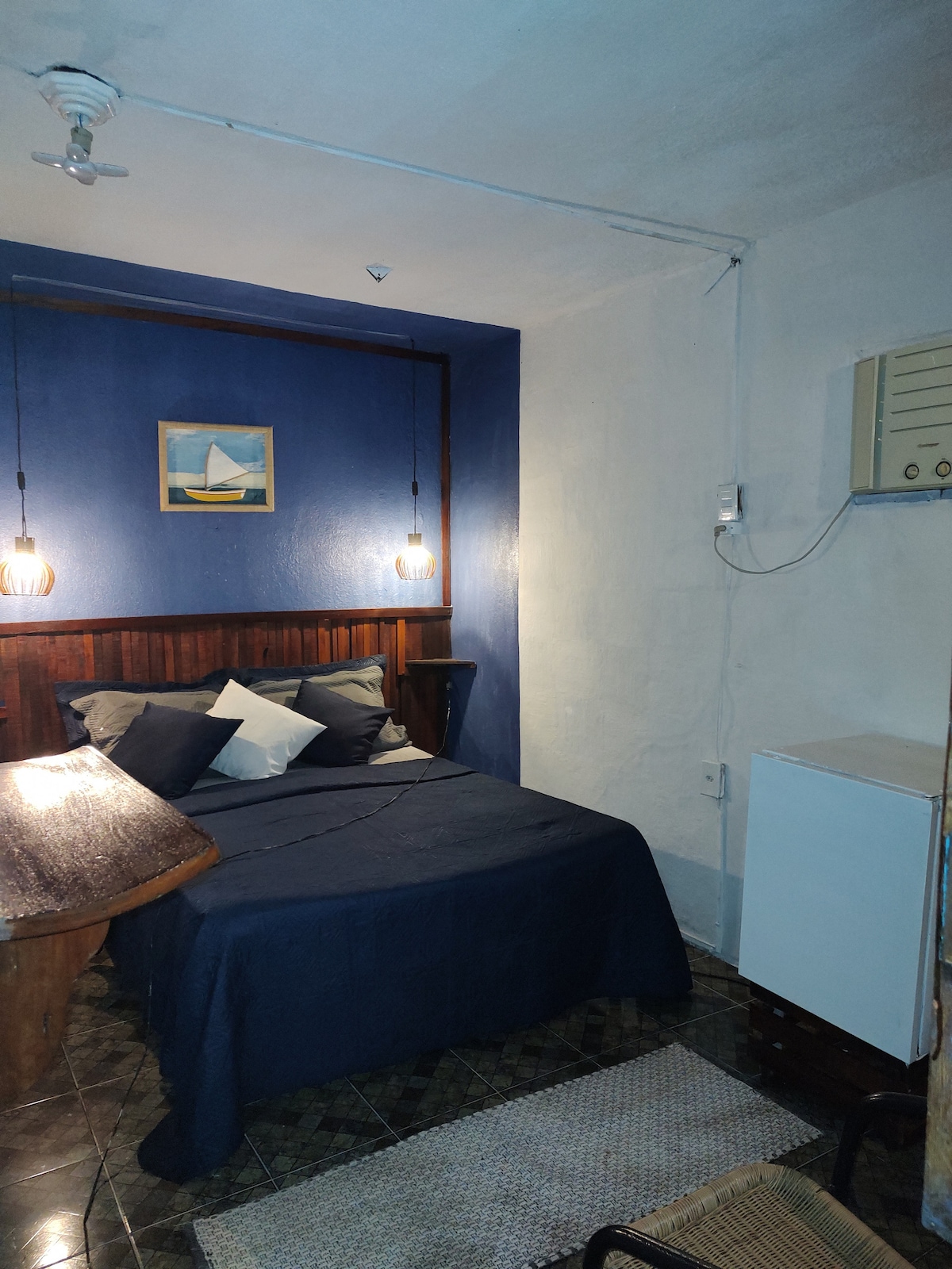 A cozy sleeping area features a spacious bed adorned with dark bedding and multiple decorative pillows. Soft lighting is provided by hanging fixtures, while a small refrigerator is positioned nearby. The walls display a calming blue hue, complemented by a piece of wall art showcasing a sailboat.