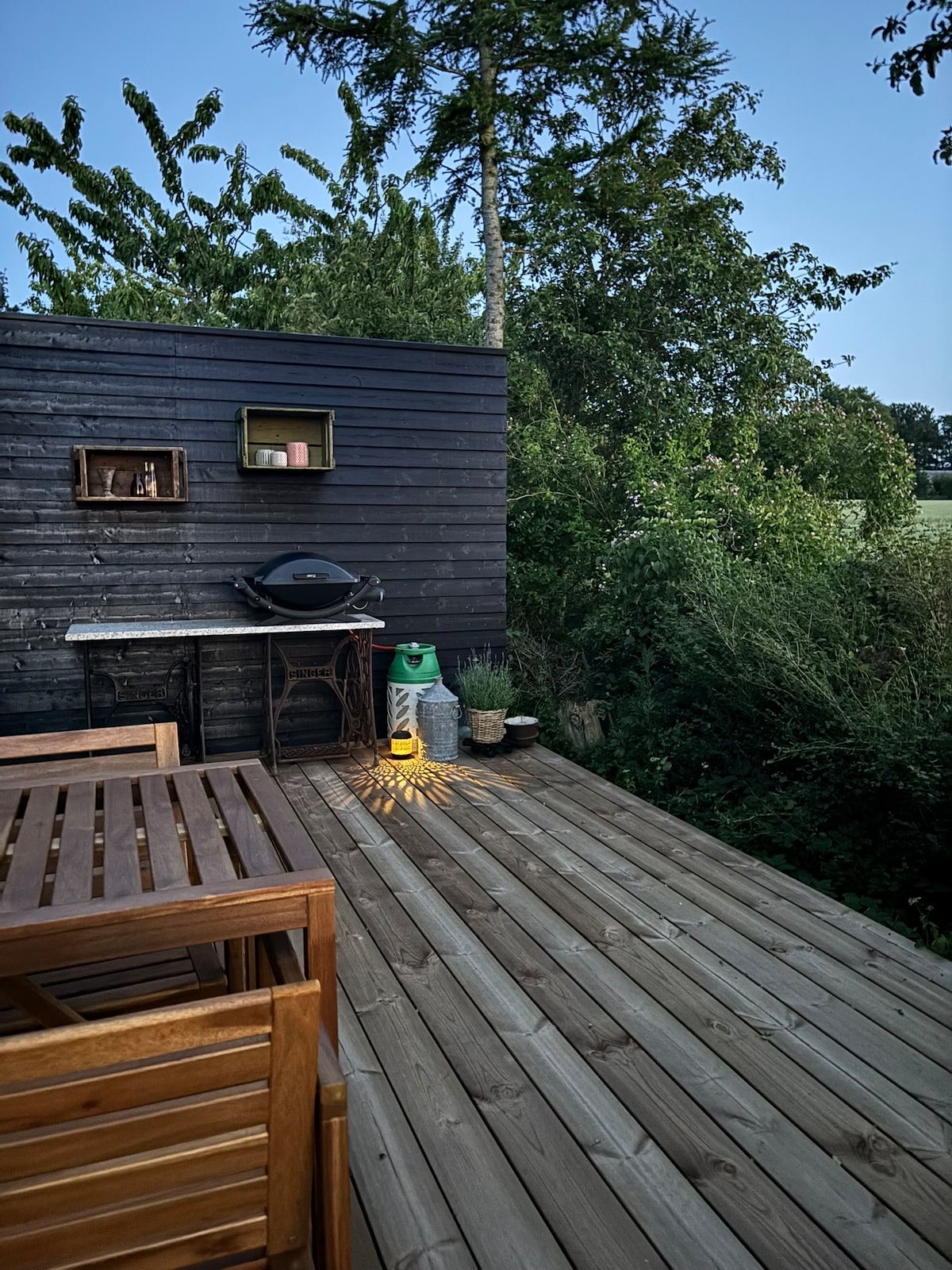 A wooden terrace is shown with a seating area made from natural wood. A small outdoor kitchen setup includes a grill and storage. Lush greenery surrounds the space, creating a serene environment. Soft lighting highlights the area, contributing to a cozy atmosphere.