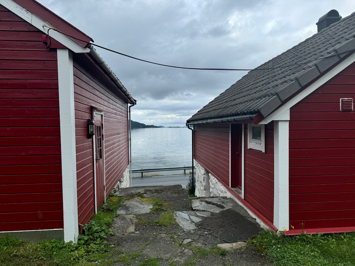 A path leads between two red wooden buildings towards a calm body of water. The ground is made of stones and grass, with gray clouds visible in the sky above. The peaceful surroundings suggest a serene atmosphere suited for relaxation.