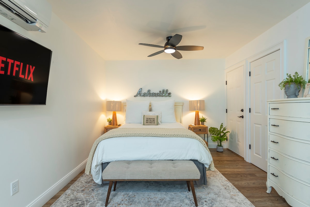 A bright and inviting studio bedroom is showcased, featuring a spacious bed dressed in soft linens. A smart TV is mounted on the wall, and a ceiling fan hangs above. Nightstands with lamps frame the bed, and a cozy area rug lies beneath a stylish bench at the foot.