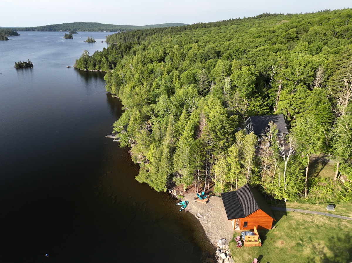 An aerial view reveals a serene landscape surrounding a cozy guest house nestled among trees. Skiff Lake lies placidly next to the shoreline, dotted with kayaks and a sandy beach area. Lush greenery and distant islands enhance the natural beauty of the setting.