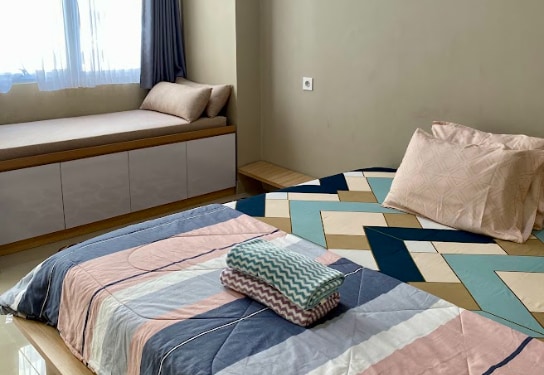 A compact sleeping area features a spacious bed covered with a patterned blanket and decorative pillows. A cozy seating nook with a cushioned bench is present by the window, with natural light filtering in and reflecting off the smooth floor.