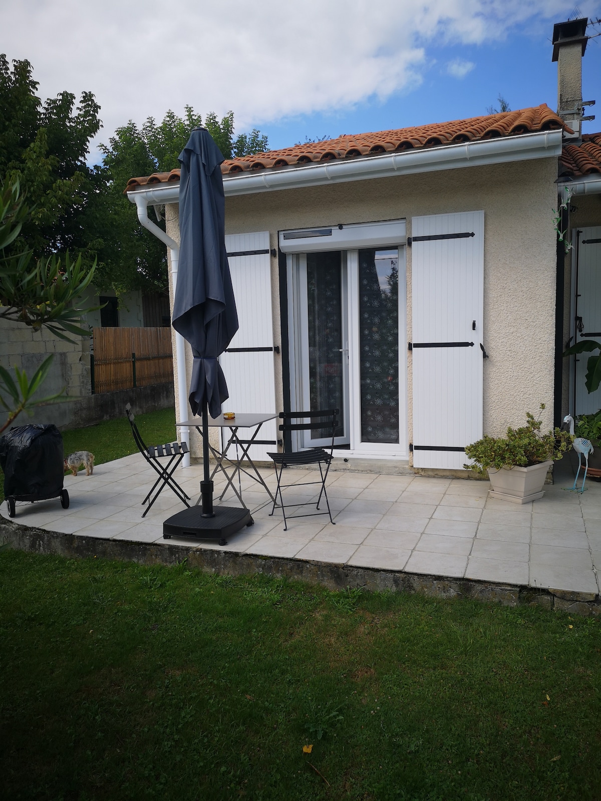 A terrace is showcased with a tiled surface, featuring a dining set that includes a table and two chairs. A large umbrella provides shade, while a charcoal barbecue is positioned nearby. Green grass surrounds the area, enhancing the outdoor space.