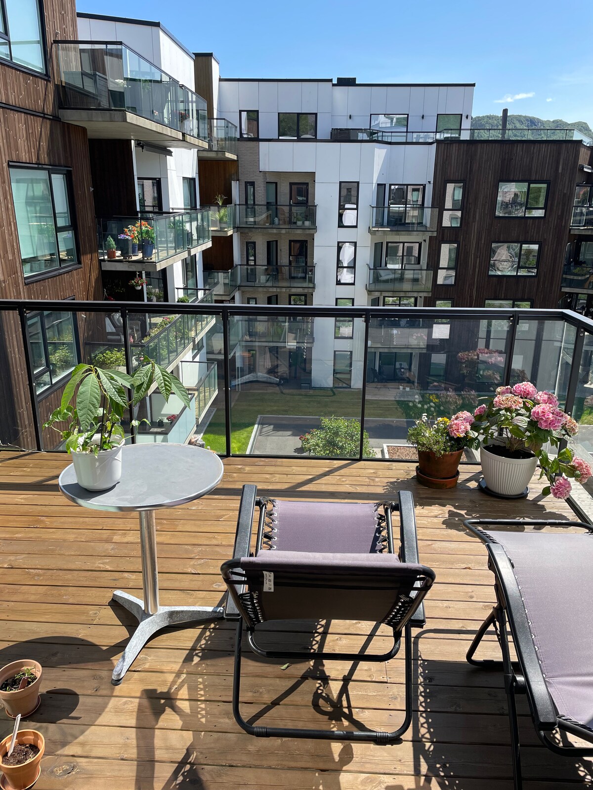 A private balcony area features a round table accompanied by a lounge chair, both positioned on wooden decking. Potted plants, including a leafy green plant and blooming flowers, add a touch of greenery. The view showcases neighboring apartment buildings with balconies and greenery.