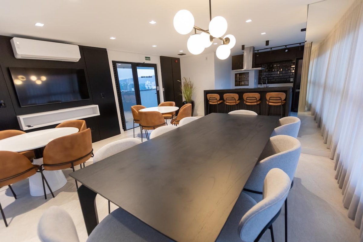 A modern dining area is equipped with a large black table surrounded by various chairs. Soft lighting illuminates the space, complemented by a contemporary chandelier. A smart TV is mounted on the wall, and an open kitchen area is visible in the background.