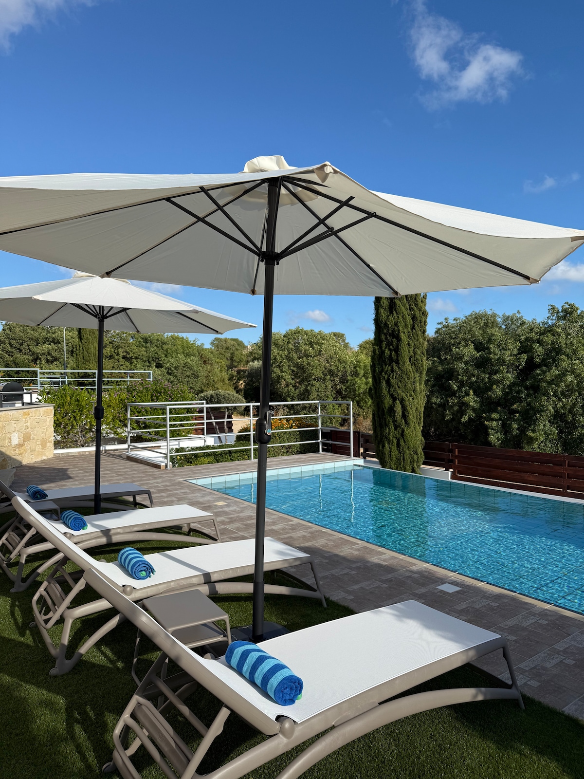 Sun loungers are arranged beside a rectangular swimming pool, each accompanied by a white umbrella. Blue and white towels are neatly placed on the loungers. Lush greenery surrounds the area, with a clear blue sky overhead.