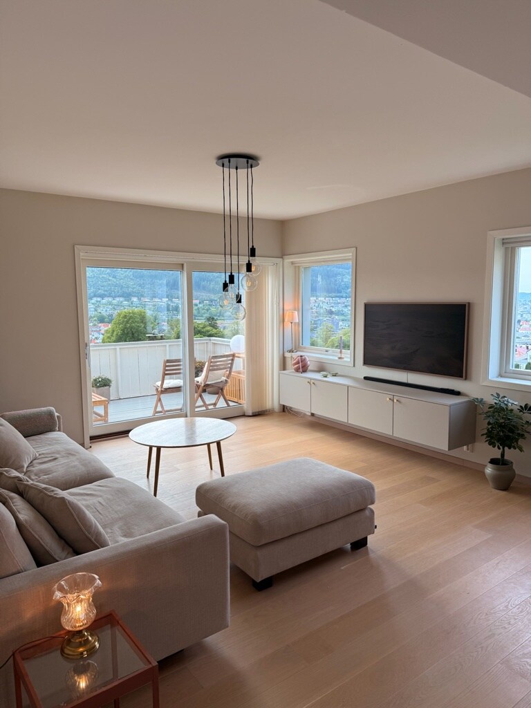 A spacious living room is illuminated by natural light, featuring a comfortable sofa and a plush ottoman. A coffee table sits in the center, with a view of the balcony visible through large sliding glass doors. A television is mounted on the wall beside a light-colored media console.