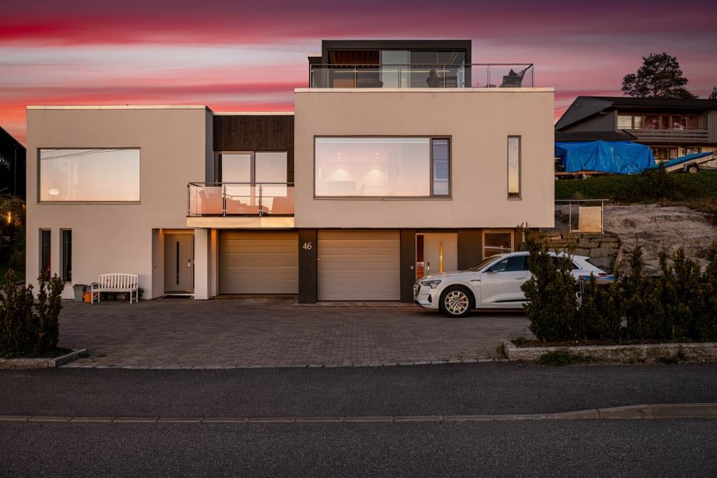 A modern two-story home is featured, presenting a sleek façade with large windows. The spacious driveway leads to two garage doors, while a white vehicle is parked nearby. A subtle landscape sets the backdrop, with a vibrant sunset sky enhancing the visual appeal.