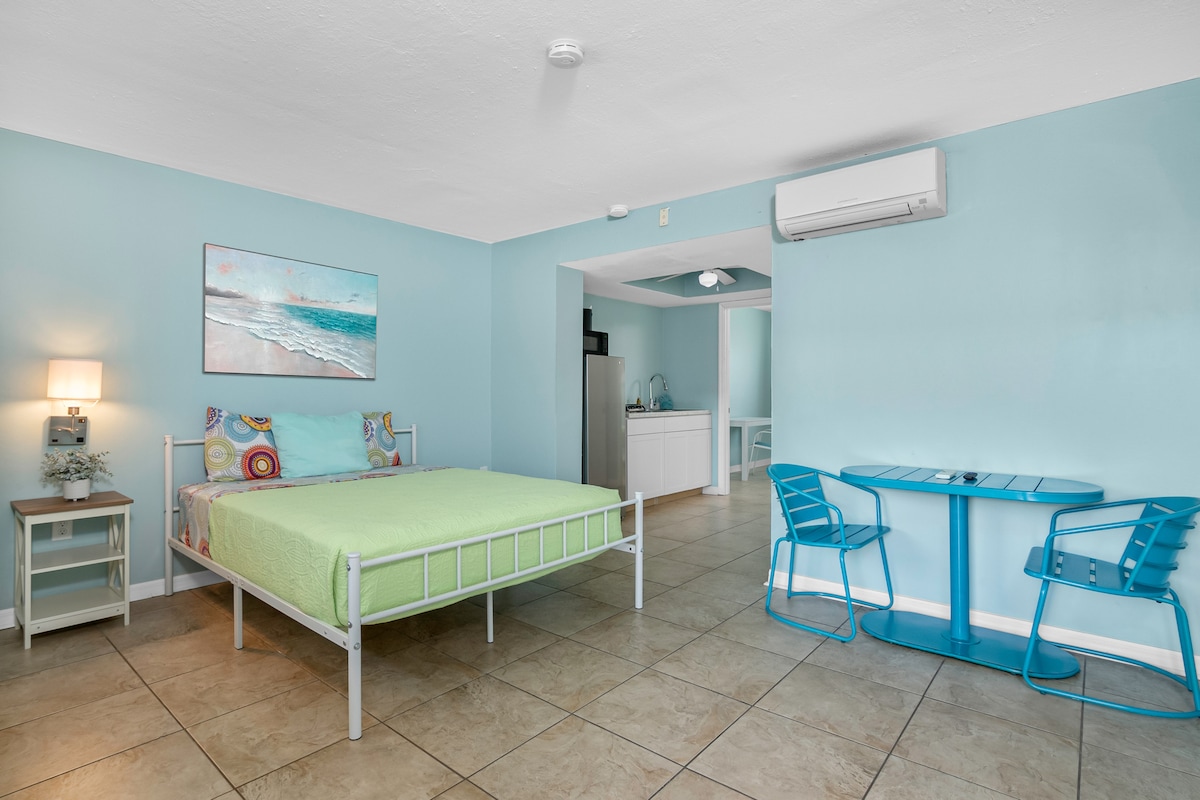 A bright room features light blue walls and a spacious bed covered with a vibrant green blanket. A small dining table with blue chairs is positioned near the kitchenette, while a piece of beach-themed artwork hangs above the bed, adding a relaxed ambiance.