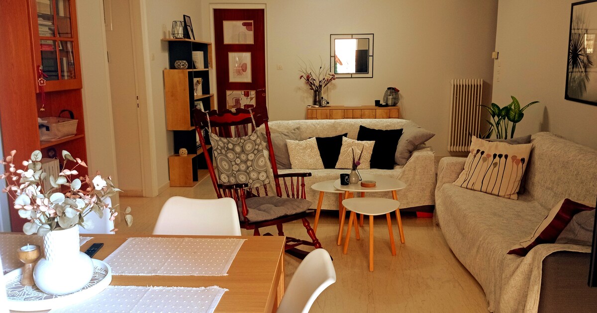 A spacious living area features a light-colored sofa adorned with decorative pillows, a cozy rocking chair, and two small coffee tables. A dining area is visible with a simple wooden table, and a bookshelf offers additional storage. Natural light brightens the room, creating a relaxed atmosphere.
