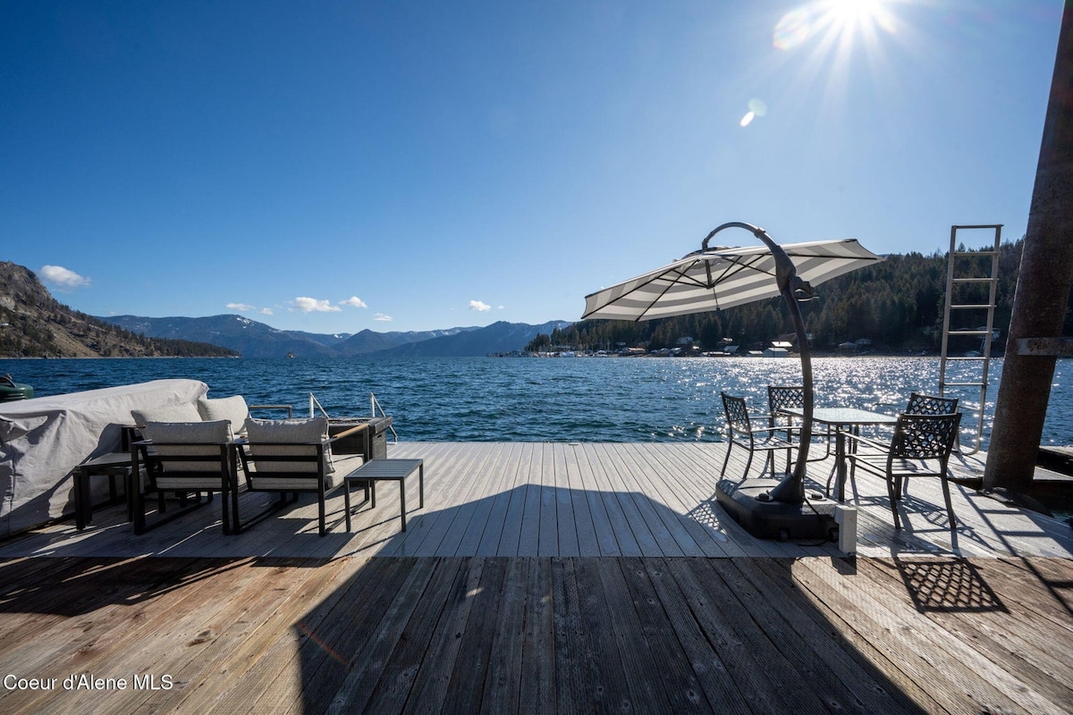 A spacious dock extends over clear waters, offering views of the surrounding mountains. Lounge chairs and a shaded seating area are visible, providing a relaxed space for guests. Sunlight reflects off the water, enhancing the serene atmosphere of the setting.