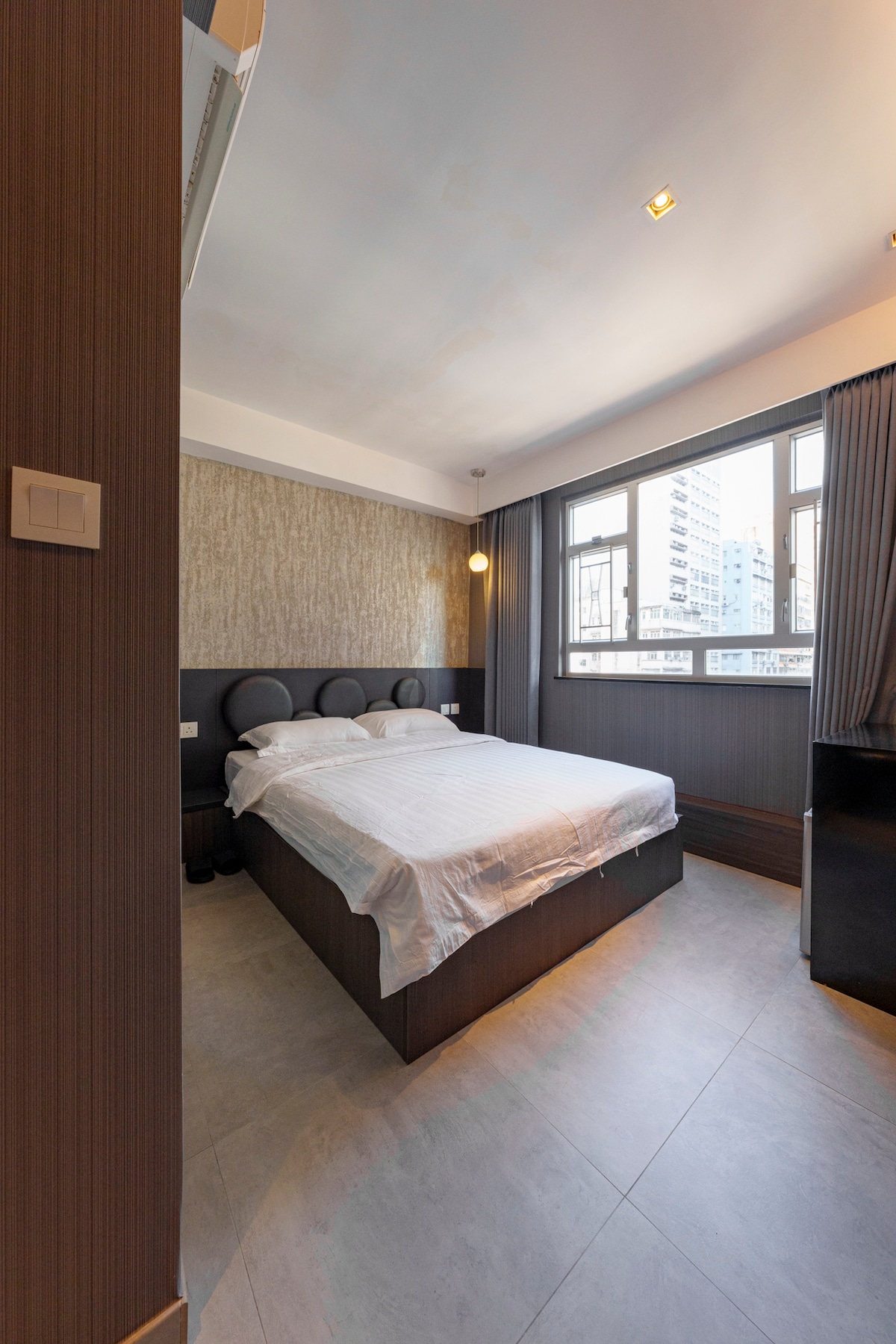 A cozy bedroom is presented with a spacious bed covered by a white linen sheet. Large windows allow natural light to enter the room, which features minimalistic design elements, including textured wall accents and dark curtains. Soft lighting is provided by a ceiling fixture.