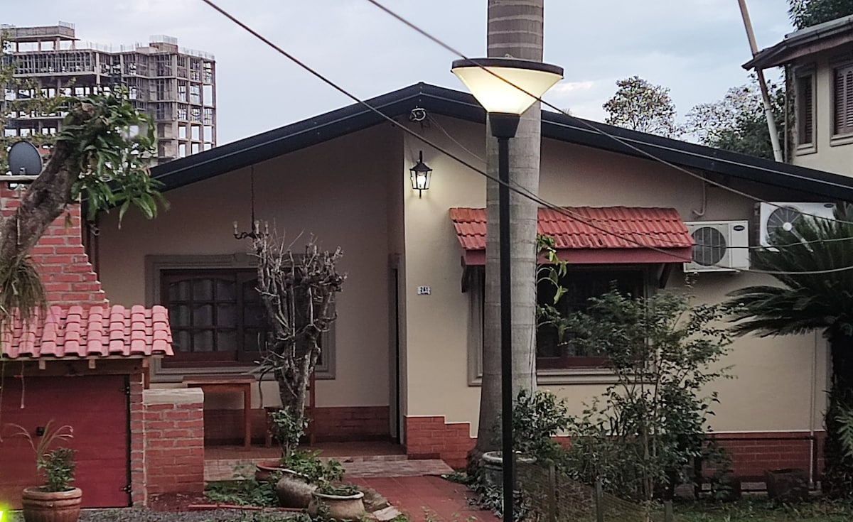 The exterior of a charming house is showcased, featuring a sloping roof and a red tile overhang. A small porch is visible, with a simple wooden chair placed beside potted plants. Soft lighting from a nearby lamp highlights the entrance amidst a backdrop of greenery.